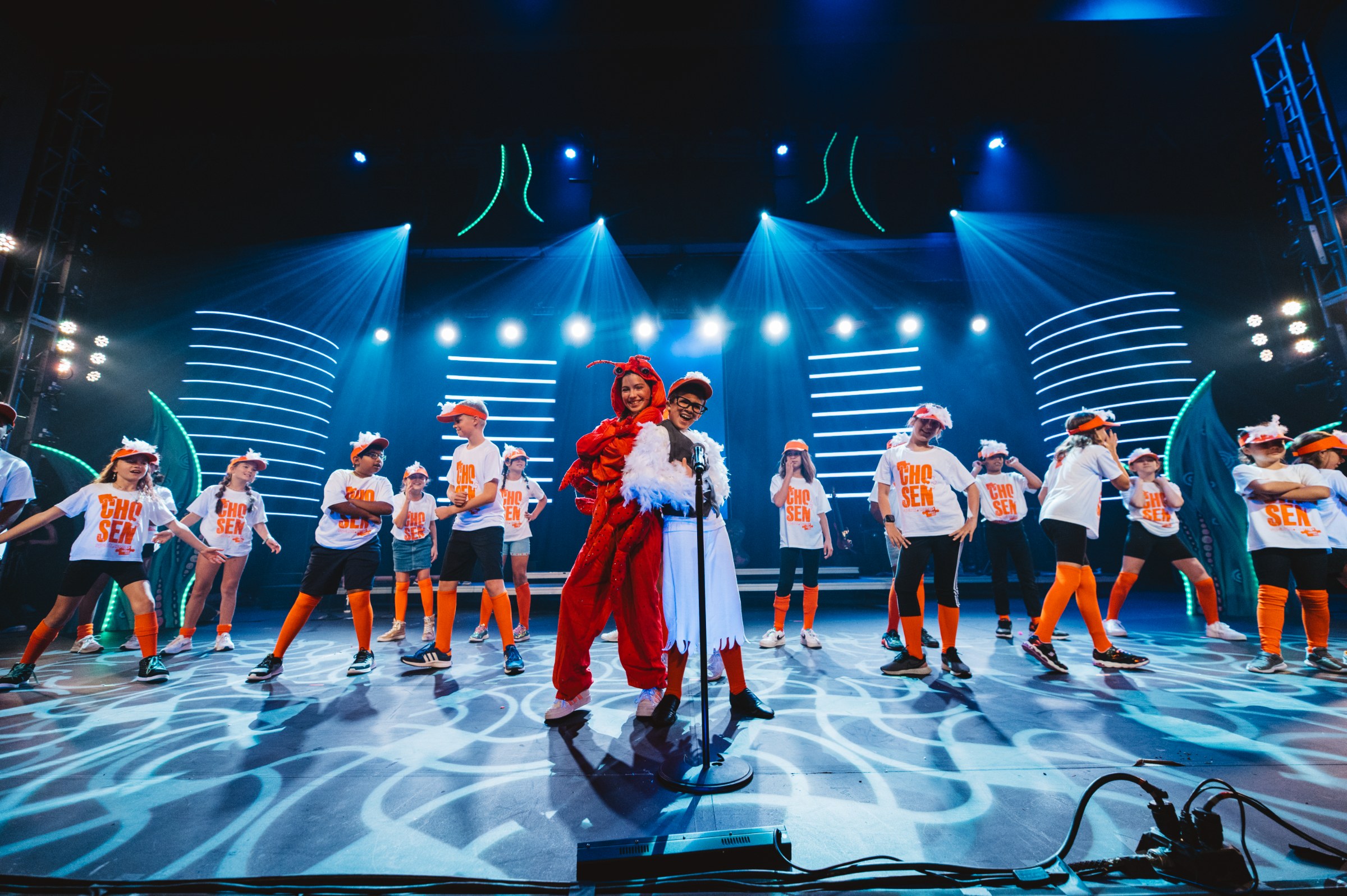 Kaleo students in orange 'Chosen' tees performing on stage with lobster and chef leads under blue lights